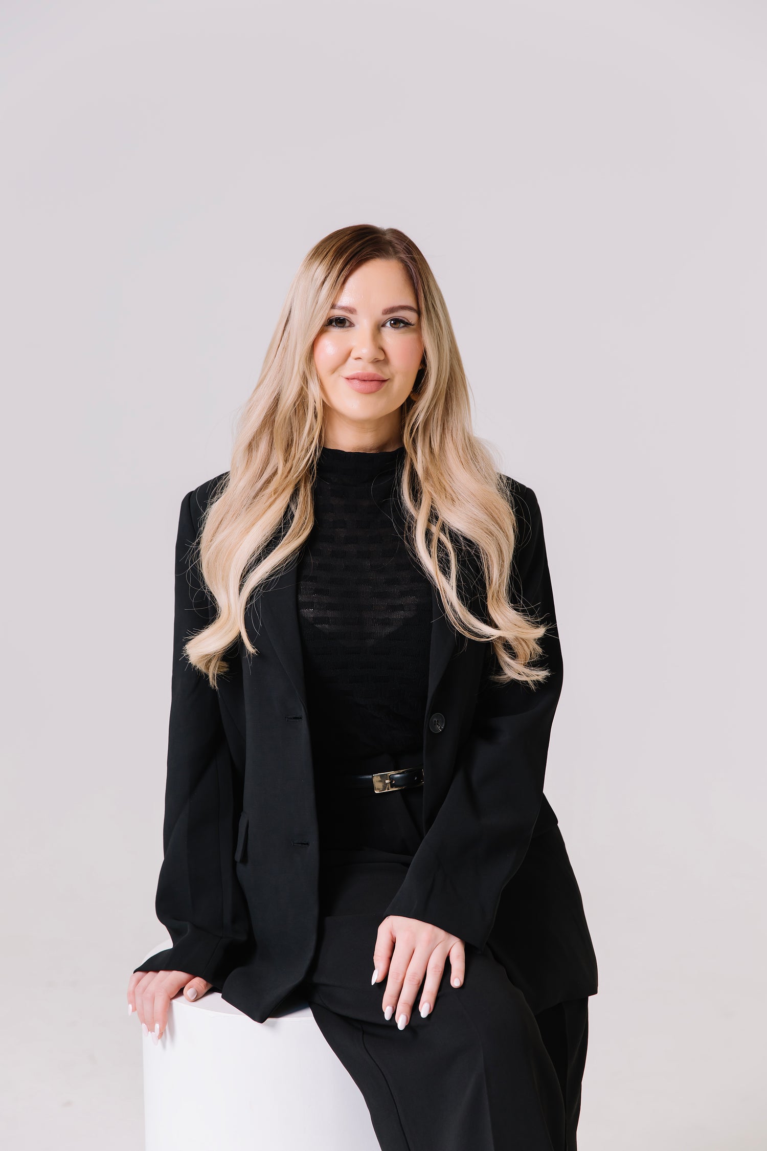 Woman in a black suit sitting on a white stool against a light gray background